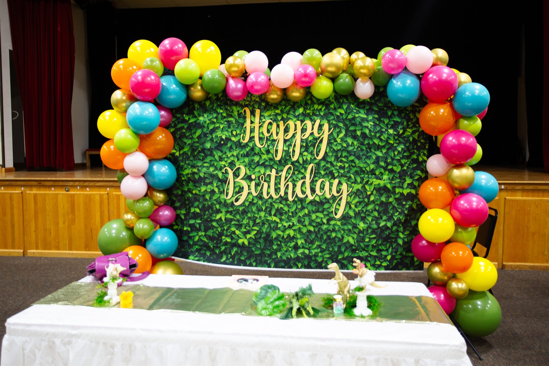 Dinosaur birthday stage with rainbow balloon arch, greenery Happy Birthday wall and gold dinosaur table