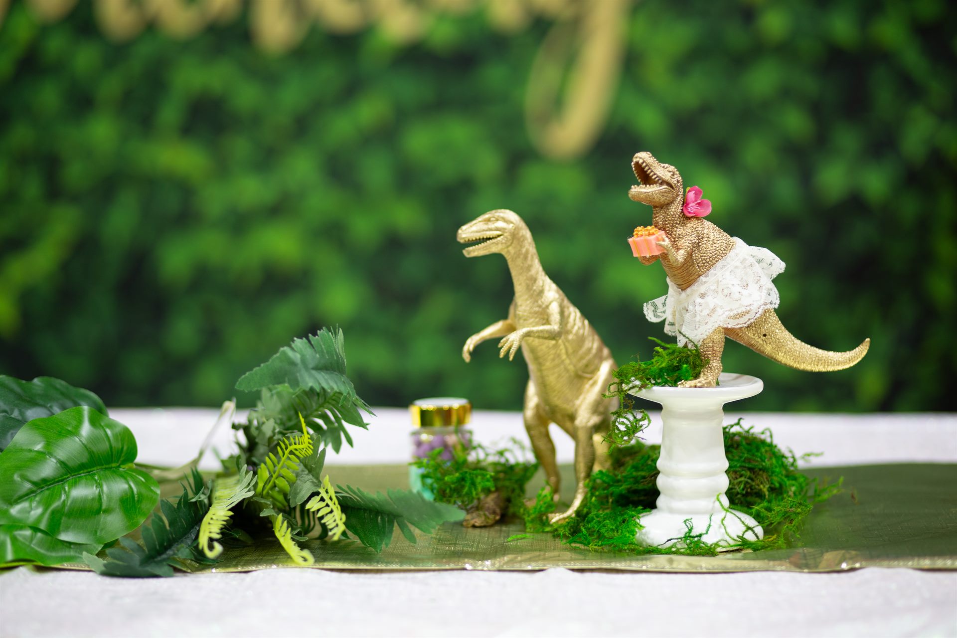 Two gold dinosaurs styled with lace, ferns and moss against a greenery wall