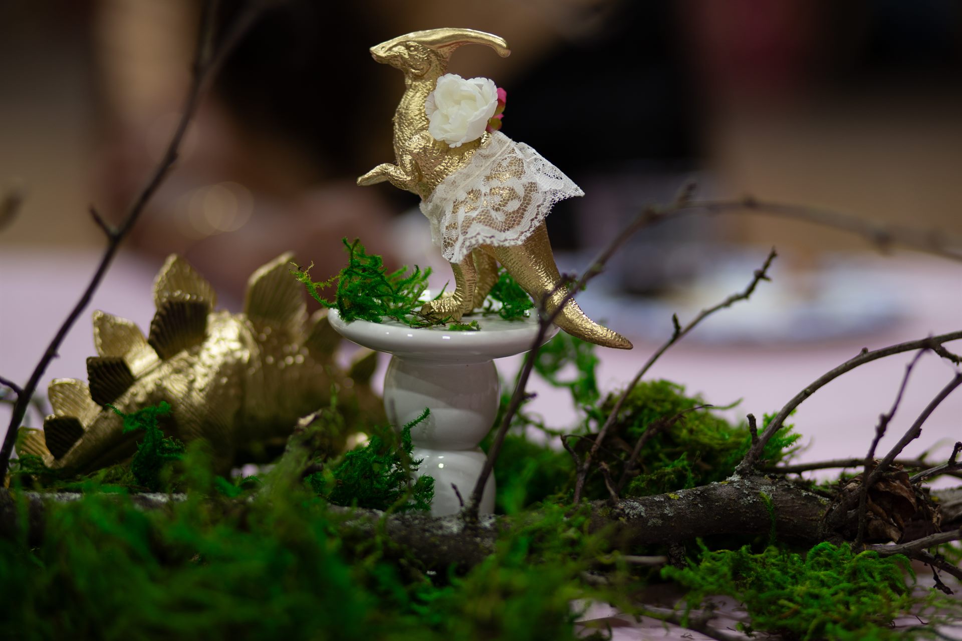 Gold dinosaur in a lace tutu on a white pedestal surrounded by moss and branches